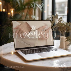 May include: A silver laptop computer with a gold trim is open on a white marble table. The laptop screen shows a website with the text "MOONICEO LACONIC CLOTHES" and a photo of a woman's hand wearing a ring. The background includes a green plant and a glass vase.
