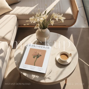 Puede incluir: Una mesa auxiliar de mármol blanco con una revista, una taza de café y un jarrón de flores blancas. La portada de la revista presenta una flor rosa con la palabra "CONSIDERED" en letras grandes.