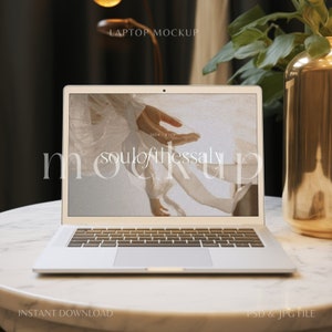 May include: A silver laptop computer with a white background on the screen. The screen shows a photo of a hand holding a piece of fabric. The text "soulofthessay" is visible on the screen. The laptop is on a white marble table.