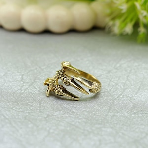 Könnte beinhalten: Ein goldfarbener Ring mit einem Krallendesign. Der Ring hat drei markante krallenartige Zinken, die sich vom Band erstrecken, mit aufwendigen Details an den Krallen und dem Band des Rings. Der Ring ist auf einer hellgrauen Oberfläche platziert.