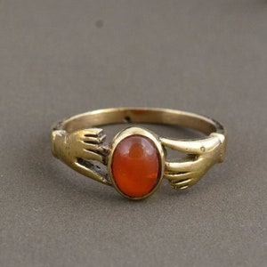 Puede incluir: Un anillo dorado con una piedra preciosa naranja ovalada en el centro. El diseño del anillo incorpora dos manos estilizadas a cada lado de la piedra preciosa, creando una joya única y simbólica.