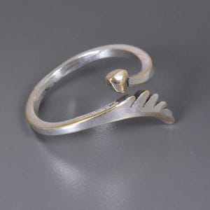 Pu&ograve; includere: Anello argentato con un dettaglio a forma di cuore e un motivo a foglia stilizzato. L'anello ha un design curvo e aperto con un bordo dorato. Il cuore &egrave; piccolo e si trova nella parte superiore dell'anello.