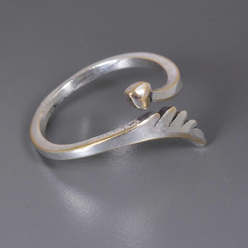 Pu&ograve; includere: Anello argentato con un dettaglio a forma di cuore e un motivo a foglia stilizzato. L'anello ha un design curvo e aperto con un bordo dorato. Il cuore &egrave; piccolo e si trova nella parte superiore dell'anello.