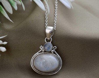 May include: A silver pendant necklace featuring a large, oval, iridescent moonstone. A smaller, round moonstone sits above the larger stone. The pendant is suspended from a delicate silver chain. The necklace is a piece of jewelry.