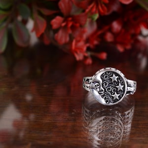 以下が含まれることがあります： 銀色のリング。三日月と三つの星が円形のフレームに収められ、渦巻き模様が施されています。暗く反射する表面に置かれています。