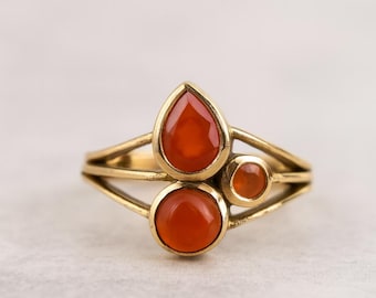 May include: A gold-toned ring featuring three orange-red gemstones. The ring has a unique design with a teardrop-shaped stone at the top and two round stones below. The gemstones are set in a delicate, open-work setting.