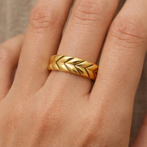 May include: A gold ring with a repeating chevron pattern. The ring is a bright, shiny gold color and has a textured surface. The design is a series of V-shaped lines that create a geometric pattern.