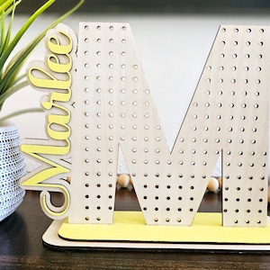 Könnte beinhalten: Ohrmanshalter aus Holz in Form des Buchstabens M mit gelben Punkten und dem Namen "Marlee" in gelben Buchstaben.