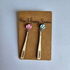 Puede incluir: Un par de pendientes de madera en forma de bate de béisbol con un dije de béisbol rosa y azul. Los pendientes están personalizados con el nombre "Emma" en cada bate. Los pendientes se muestran en una tarjeta marrón con el texto "Five Happy Swans".
