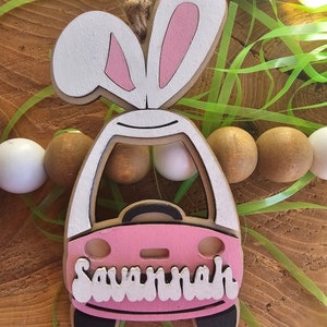 May include: Pink wooden car with bunny ears and the name Savannah written in white on the front. The car is decorated with black details and white accents.