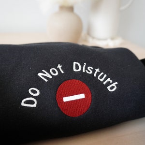 May include: Black sweatshirt with the embroidered phrase "Do Not Disturb" in white, above a red circle with a white horizontal line. The sweatshirt is folded, with the text prominently displayed.