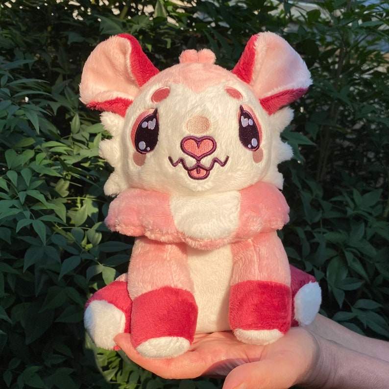 May include: A plush toy in the shape of a small animal with pink and red fur. It has large eyes, a pink nose, and a small mouth. The toy is sitting on a hand.