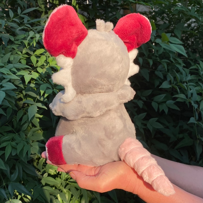 May include: A plush toy in the shape of a gray rat with red ears and a pink tail. The rat is wearing a white hooded jacket.