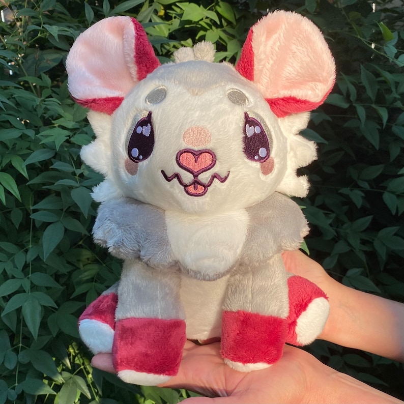 May include: A plush toy shaped like a mouse with large pink ears, a white face with embroidered eyes, a pink nose, and a pink heart on its chest. The toy is wearing red and gray pants and has a white fluffy tail.