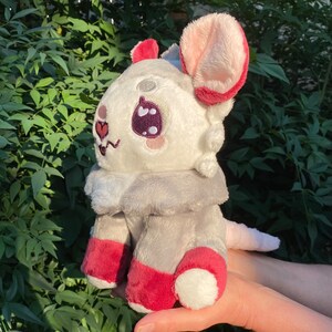 May include: A plush toy shaped like a small animal with white fur, red ears, and red accents on its feet. The toy has large, black eyes and a pink nose.