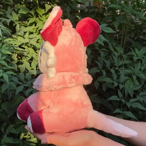 May include: Pink plush toy with a white face and red ears. The toy is sitting on a hand and has a long tail.