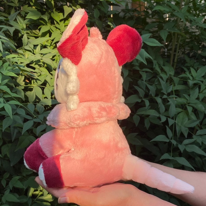 May include: Pink plush toy with a white face and red ears. The toy is sitting on a hand and has a long tail.