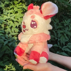 May include: Pink and white plush toy with a heart on its face and a pink and white tail. The toy has large ears and is sitting on a hand.