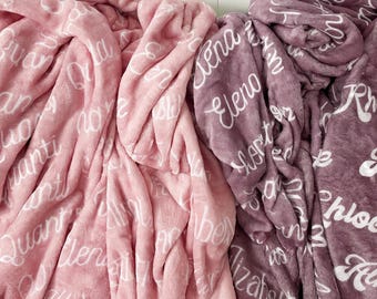 May include: Two plush blankets in shades of pink and purple. Each blanket is covered in white script names. The blankets are folded and draped, showing the soft texture of the fabric. The background is a white, slatted wall.