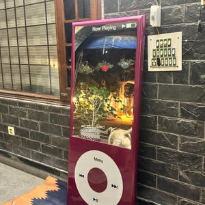May include: A large, magenta, rectangular display screen shaped like a classic digital music player. The screen shows a Now Playing interface with a garden scene and the text Find Yourself. Below the screen is a white circular control wheel.