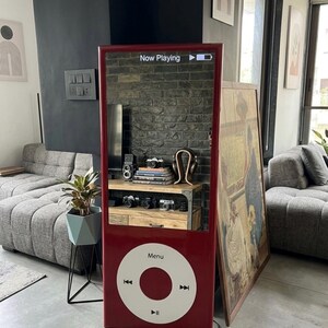 May include: A tall, rectangular, red floor mirror designed to resemble a classic portable music player. The top section is a reflective mirror surface with the text "Now Playing" and a battery icon. The bottom features a large white circular control wheel.