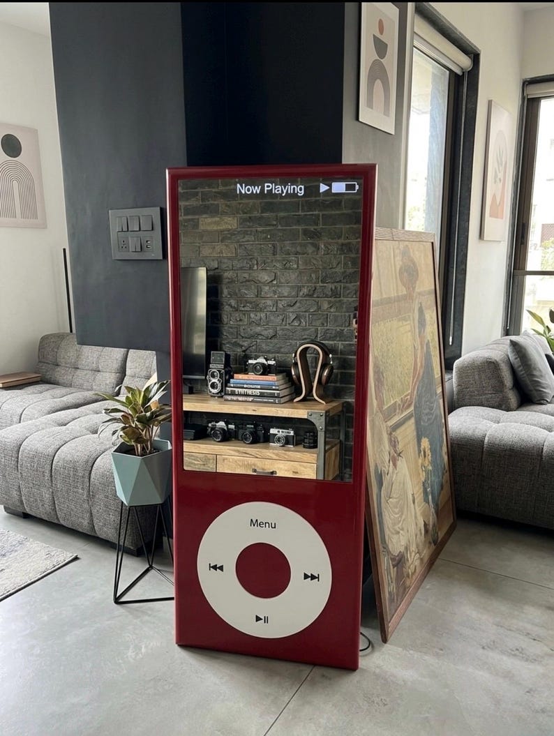 May include: A tall, rectangular, red floor mirror designed to resemble a classic portable music player. The top section is a reflective mirror surface with the text "Now Playing" and a battery icon. The bottom features a large white circular control wheel.