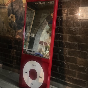 May include: A tall, red, rectangular floor mirror designed to resemble a classic digital music player. The top section features a reflective glass surface with the text Now Playing, while the bottom section displays a large white circular control wheel.