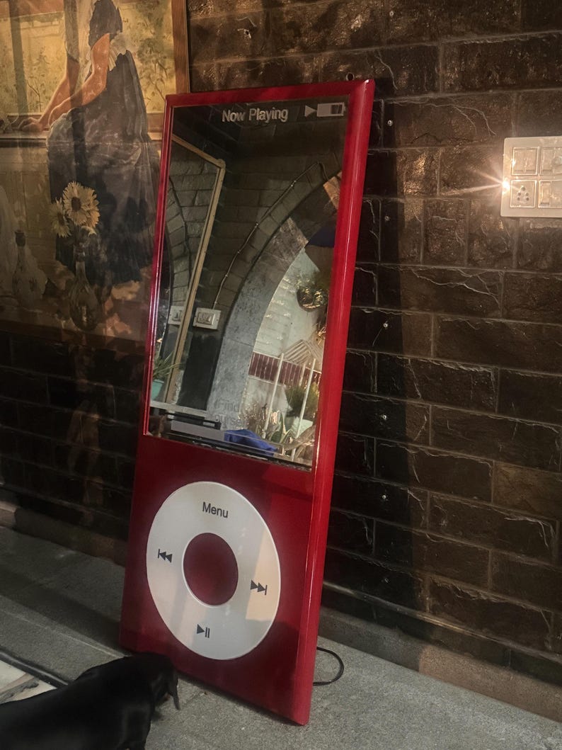 May include: A tall, red, rectangular floor mirror designed to resemble a classic digital music player. The top section features a reflective glass surface with the text Now Playing, while the bottom section displays a large white circular control wheel.