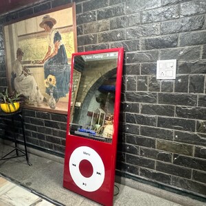 May include: A tall, rectangular, red floor mirror designed to resemble a vintage digital music player. It features a large, white circular click wheel with a central button and playback icons, and a top screen area displaying "Now Playing".