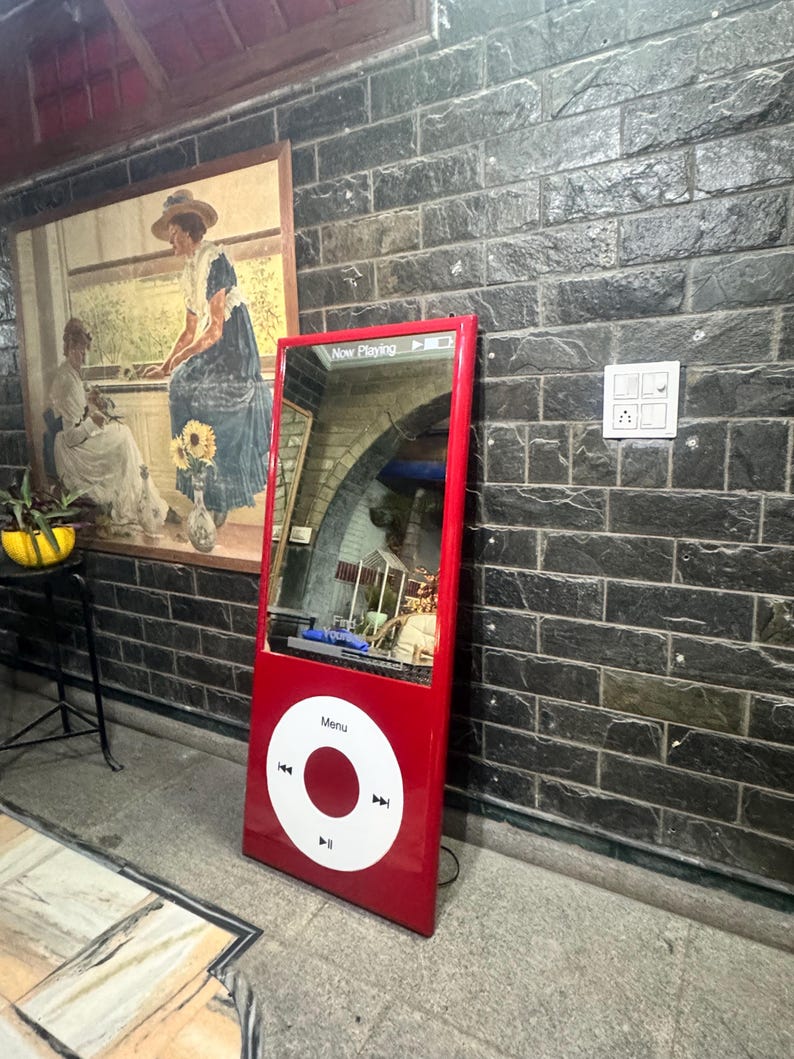 May include: A tall, rectangular, red floor mirror designed to resemble a vintage digital music player. It features a large, white circular click wheel with a central button and playback icons, and a top screen area displaying "Now Playing".