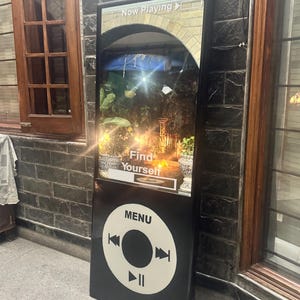 May include: A tall, black, rectangular sign shaped like a vintage digital music player. The top section displays a screen with the text "Now Playing" and "Find Yourself" over a garden scene. The bottom section features a white circular menu interface.