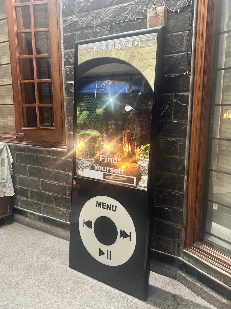 May include: A tall, black, rectangular sign shaped like a vintage digital music player. The top section displays a screen with the text "Now Playing" and "Find Yourself" over a garden scene. The bottom section features a white circular menu interface.