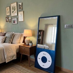 May include: A tall, rectangular floor mirror designed to resemble a classic digital music player. The frame is dark blue with a large white circular control wheel at the bottom featuring the text Menu and playback icons. The screen area is reflective.
