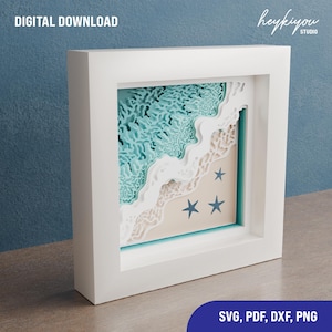 Könnte beinhalten: Ein weißer Rahmen mit einem mehrschichtigen Papierschnitt-Design von Ozeanwellen und einem Sandstrand mit blauen Seesternen. Das Design ist in Blautönen, Weiß und Beige gehalten. Der Rahmen befindet sich auf einer Holzoberfläche. SVG, PDF, DXF, PNG.