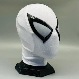 May include: A white Spider-Man mask with black-outlined eye lenses. The mask has a textured surface and is displayed on a black stand. The eye lenses have a grid-like pattern.