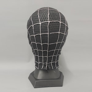 Puede incluir: M&aacute;scara de spandex negra y blanca con un dise&ntilde;o de telara&ntilde;a. La m&aacute;scara est&aacute; en un soporte negro.