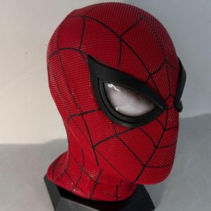 Op de afbeelding: Een rood en zwart Spider-Man masker met een getextureerd oppervlak en zwarte oogframes. Het masker heeft een webpatroon en wordt weergegeven op een zwarte standaard. De ooglenzen zijn wit met een mesh-ontwerp.