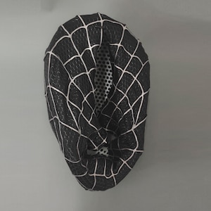 Puede incluir: M&aacute;scara de tela negra y blanca con una telara&ntilde;a. La m&aacute;scara est&aacute; hecha de un tejido el&aacute;stico y tiene un agujero para los ojos.