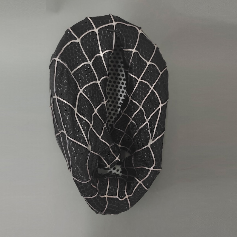 Puede incluir: M&aacute;scara de tela negra y blanca con una telara&ntilde;a. La m&aacute;scara est&aacute; hecha de un tejido el&aacute;stico y tiene un agujero para los ojos.