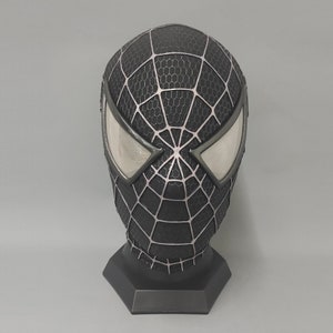 Puede incluir: M&aacute;scara de Spider-Man negra y plateada con un dise&ntilde;o de telara&ntilde;a. La m&aacute;scara est&aacute; sobre un soporte negro.