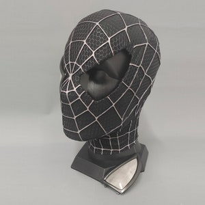 Puede incluir: M&aacute;scara de Spider-Man en blanco y negro con un dise&ntilde;o de telara&ntilde;a detallado. La m&aacute;scara se muestra en un soporte negro.