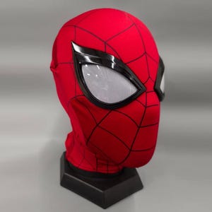 May include: A red and black Spider-Man mask with a mesh eye covering. The mask is on a black stand.