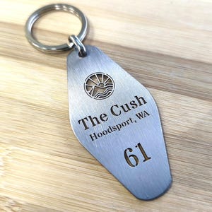 May include: A brushed metal keychain with a circular ring. The tag has the text "The Cush Hoodsport, WA 61" and a sun and water logo. The keychain is on a light wood surface.