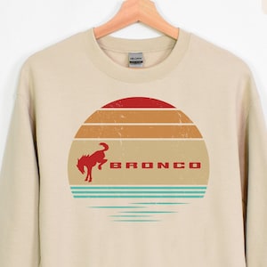 May include: Sand-colored sweatshirt with a retro design featuring a red bucking bronco inside a sunset graphic. The word "BRONCO" is printed in red. The sweatshirt is hanging on a wooden hanger.