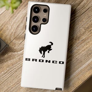 Könnte beinhalten: Eine weiße Handyhülle mit einer schwarzen Pferdesilhouette und dem Wort "BRONCO". Die Hülle hat ein Kameramodul mit mehreren Objektiven. Die Hülle liegt auf einer Holzoberfläche.