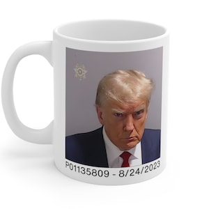 May include: White ceramic mug with a photo of a man with blonde hair and a red tie. The photo is a mugshot with the text "P01135809 - 8/24/2023" at the bottom.