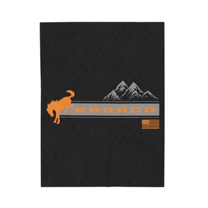 Puede incluir: Manta negra con un gráfico de bronco naranja, la palabra "BRONCO" en naranja y una pequeña bandera estadounidense. El diseño incluye una cordillera en gris y blanco, sobre un patrón de diamantes.