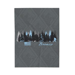 Peut inclure: Un fond gris avec un motif répétitif de montagnes en forme de losange. Une rangée d'arbres bleus et noirs avec une silhouette bleue de Bigfoot marchant vers la droite. Un petit drapeau américain se trouve sous les arbres. Le texte "Bronco" est écrit en cursive bleue sous le Bigfoot.