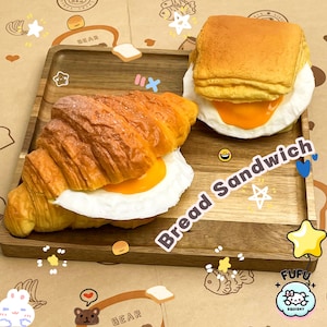 May include: Two realistic-looking food items on a wooden tray. A croissant with a fried egg and a bread sandwich with a fried egg. The tray has a brown wood grain pattern and is decorated with cartoon drawings. The text "Bread Sandwich" is written in white on the tray.
