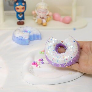 May include: A purple and white donut with iridescent sprinkles, held by a hand. The donut has a bite taken out of it, revealing a purple interior. Other decorative items are in the background, including a blue donut slice.